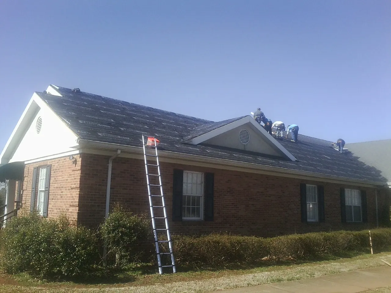 Crew tearing shingles off a brick ranch roof with a ladder set — Metal Roof Repair work by our crew in Bigfork